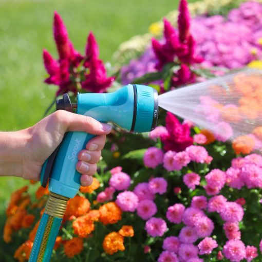 A person's hand holds a blue hose nozzle, spraying water on a vibrant garden with pink, orange, and purple flowers on a sunny day.