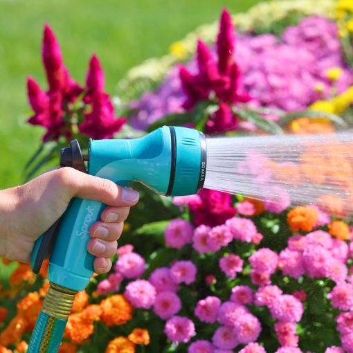 A person's hand is holding a blue garden hose with a sprayer, watering vibrant pink and purple flowers in a sunlit garden.