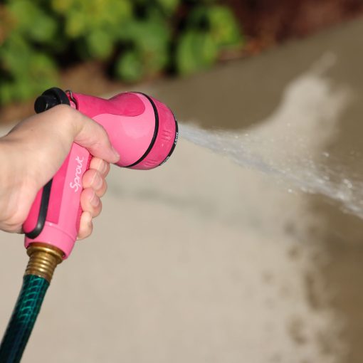 A person's hand is holding a pink garden hose nozzle, spraying water onto a concrete surface, with green foliage subtly noticeable in the background.