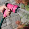 A person's hand is holding a pink garden hose nozzle, spraying water, against a blurred background of stone blocks and plants.