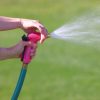 A person is holding a pink garden hose nozzle, spraying water onto a lush green lawn on a sunny day, possibly engaging in garden maintenance.