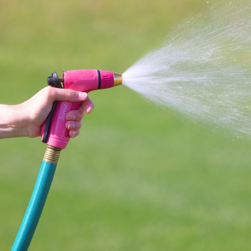 A person's hand is holding a pink garden hose nozzle, spraying a mist of water onto a sunlit green grassy lawn.