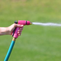 A person's hand is gripping a pink garden hose nozzle, spraying water onto green grass. It's sunny, and the background is softly focused.