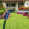 A person's hand is holding a blue hose nozzle, spraying water onto a lush green lawn with colorful flowerbeds in front of a white house.