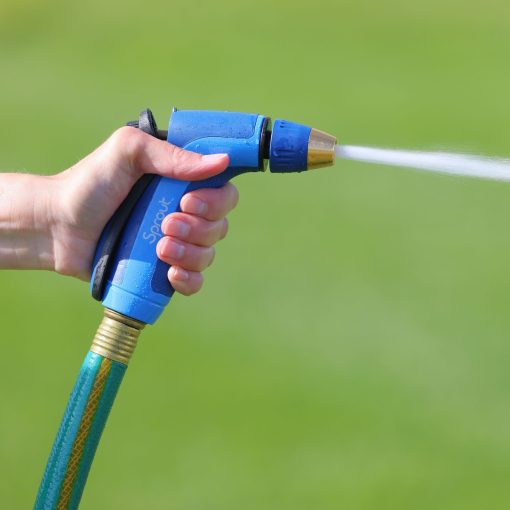 A person's hand holding a blue and black garden hose nozzle, spraying a jet of water, against a blurred green grass background.