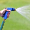 A person's hand is holding a blue garden hose with a nozzle, spraying water onto a bright green lawn under clear daylight.