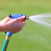 A person's hand is holding a blue and black hose nozzle, spraying water onto a green grassy lawn, with a focused jet of water visible.