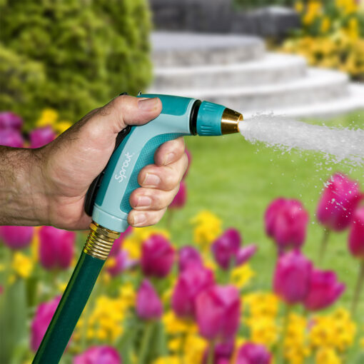 A person is holding a teal and black hose nozzle, spraying water in a vibrant garden with an abundance of pink tulips and a fountain.