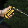 A person's hand grips a yellow and black garden hose nozzle, spraying water, with greenery blurred in the background. Focused and clear outdoor shot.