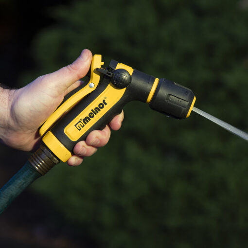 A person's hand grips a yellow and black garden hose nozzle, spraying water, with greenery blurred in the background. Focused and clear outdoor shot.