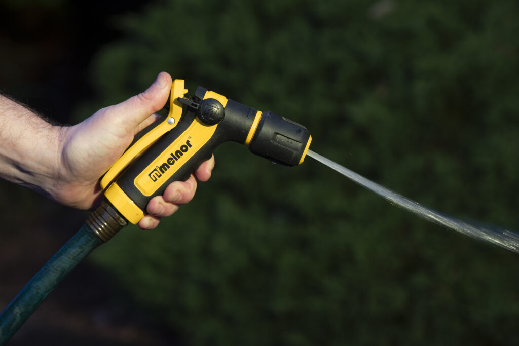 A person's hand is holding a yellow and black garden hose nozzle, spraying a jet of water, with green foliage blurred in the background.