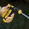 A person is holding a yellow and black garden hose nozzle emitting a strong, focused stream of water, set against a blurred green background.