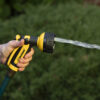 A person is holding a yellow and black garden hose nozzle, emitting a steady stream of water, with green foliage softly focused in the background.