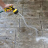 A person is cleaning a tiled surface with a high-pressure water spray nozzle, effectively removing dirt from the wet floor. The nozzle is yellow and black.