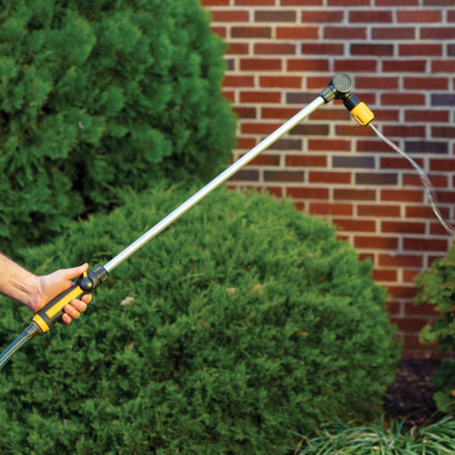 A person's hand is holding a garden hose with a long attachment, spraying water on green bushes in front of a brick wall.