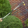 A person's hand is holding an extended water hose nozzle against a background of a brick wall and green shrubbery. The device is likely used for gardening.