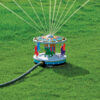 A colorful, carousel-shaped sprinkler is set on a green lawn, connected to a hose, with water jets spraying outward from its top in multiple directions.