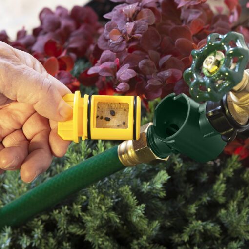 A person's hand is holding a yellow and black garden hose filter attached to a green hose with a water sprinkler head, amidst floral foliage.