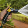 A person's hand holds a black and yellow garden hose nozzle, spraying water forcefully against a blurred green and wooden deck background.