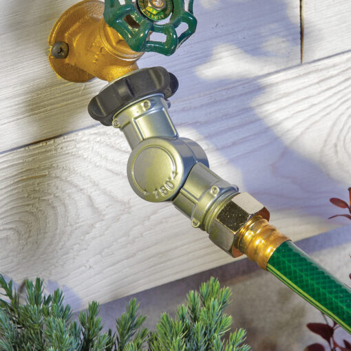 An outdoor water spigot is connected to a garden hose with a metallic quick connector. It's mounted on a wooden wall, partially shaded, with foliage below.