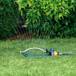 An oscillating sprinkler is watering a lush green lawn against a backdrop of dense shrubbery. It's connected to a hose, with water jets in mid-arc.