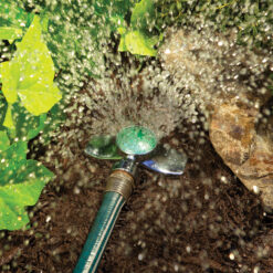 A garden hose with a sprinkler attachment is watering plants, releasing a fine spray of water droplets in the sunlight against a backdrop of greenery.