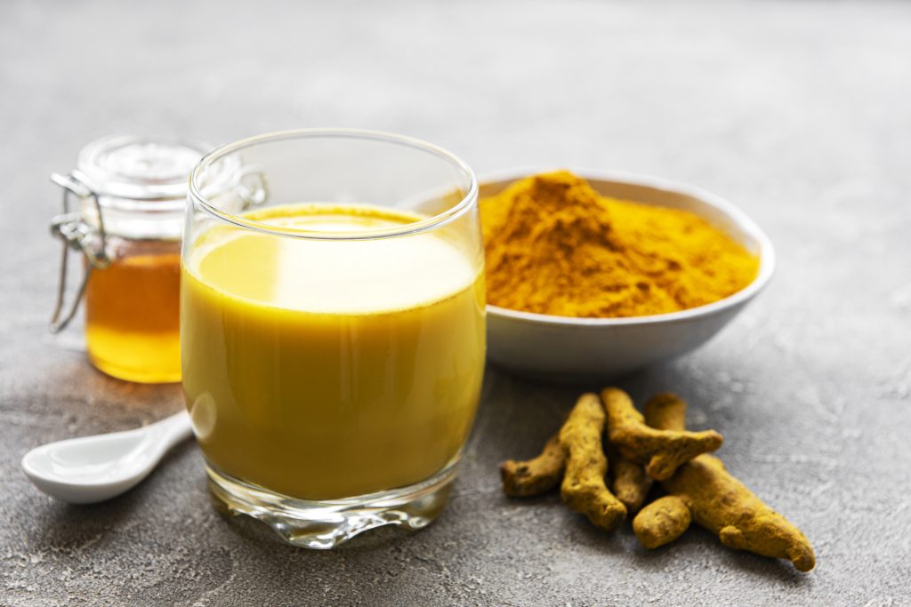 A glass of golden milk is in the foreground with turmeric powder in a bowl and honey jar behind it, accompanied by raw turmeric roots on a grey surface.