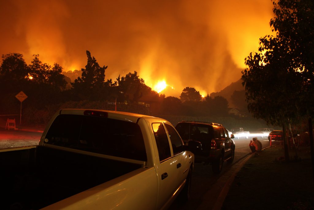 The image shows vehicles parked on a street at night while a large, intense wildfire burns in the background, illuminating the sky with an orange glow.