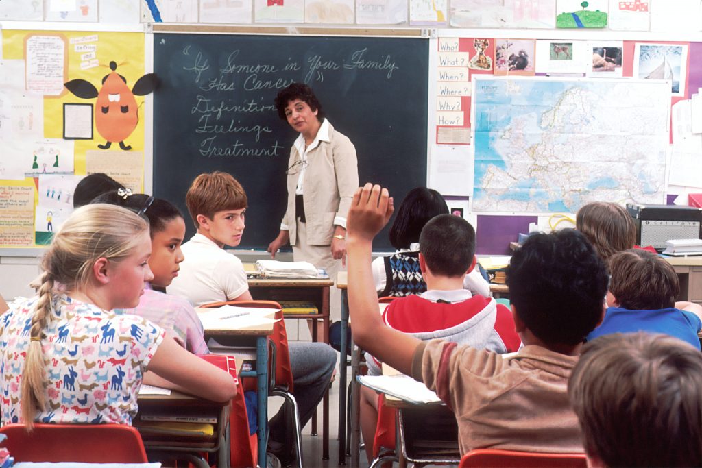 In a colorful classroom, students sit at desks, looking towards a person by the blackboard. One child raises a hand, possibly to ask a question.