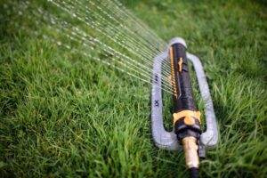 An oscillating sprinkler is watering green grass. Its black and orange body sits on a gray base connected to a hose, with water spraying out.