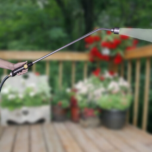 In the image, a person's hand is holding a garden hose with a nozzle, spraying water, with blooming plants and a blurred green background.