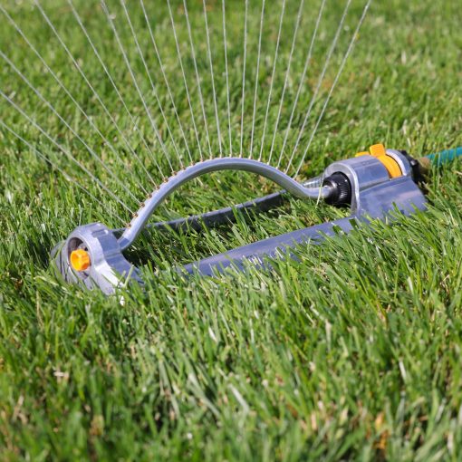 An oscillating sprinkler with a blue body and yellow fittings is lying on vibrant green grass, likely set up for watering a lawn.