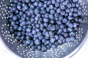 This image shows a plethora of fresh blueberries inside a metal colander with round perforations, likely being washed or drained. The background is white.