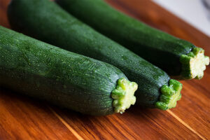 Three fresh green zucchinis with water droplets are arranged on a wooden cutting board, showing vibrant colors and healthy, intact stems.