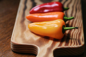 Three vibrant bell peppers—one red, two orange—arranged on a wooden cutting board with noticeable grain, photographed with a shallow depth of field.