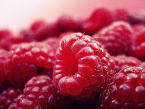 This image shows a close-up of ripe, red raspberries with a noticeable texture, set against a soft-focus background in a roseate color palette.