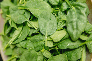 Fresh, vibrant green spinach leaves with visible water droplets, close-up, filling the frame. The leaves display a healthy, leafy texture, suggesting a raw, nutritious vegetable.