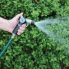 A person's hand is holding a garden hose with a nozzle, spraying water in a fine mist over a lush green hedge in the background.