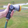 A person is holding a garden hose nozzle, ready to spray water. The nozzle is attached to a green and blue hose, set against a grassy background.