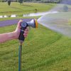 A person's hand is holding a garden hose with a spray nozzle, which is emitting a fine mist of water onto a green lawn near a lake.