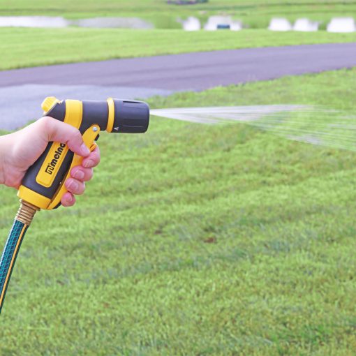 A person's hand is holding a yellow and black garden hose nozzle, spraying water onto a green lawn with a pathway in the background.