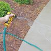 A person's hand is holding a yellow and black garden hose nozzle, spraying water onto a concrete path surrounded by greenery and mulch.